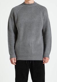 Suéter gris de punto con textura acanalada, escote redondo y mangas raglán. El ajuste es holgado con un dobladillo largo. Combinado con pantalones de pana negros.