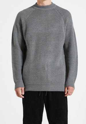 Suéter gris de punto con textura acanalada, escote redondo y mangas raglán. El ajuste es holgado con un dobladillo largo. Combinado con pantalones de pana negros.