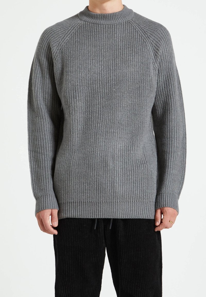 Suéter gris de punto con textura acanalada, escote redondo y mangas raglán. El ajuste es holgado con un dobladillo largo. Combinado con pantalones de pana negros.