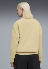 Gul sweatshirt med løs pasform, ribbede manchetter og rund halsudskæring, vist fra ryggen mod en grå baggrund.