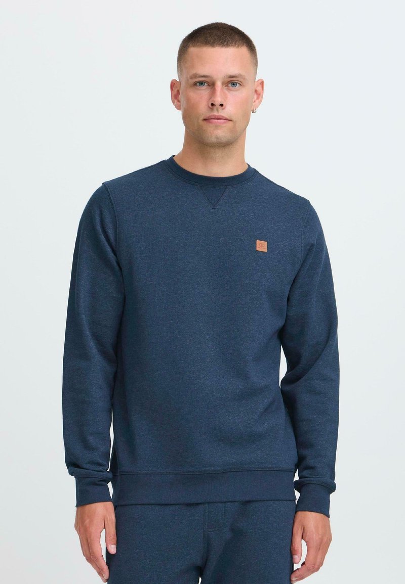 Uomo che indossa un maglione girocollo blu navy e pantaloni abbinati, in piedi contro uno sfondo chiaro uniforme, rivolto in avanti.