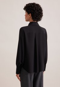 Schwarzbluse mit sanftem Fall, klassischem Kragen, langen Ärmeln und Knopfmanschetten. Glatte Textur mit lockerem Schnitt.