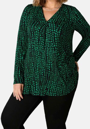 Vrouw die een zwart shirt met lange mouwen draagt met een groen onregelmatig stippenpatroon, zwarte broek en zilveren ketting, staand met één hand op de heup.