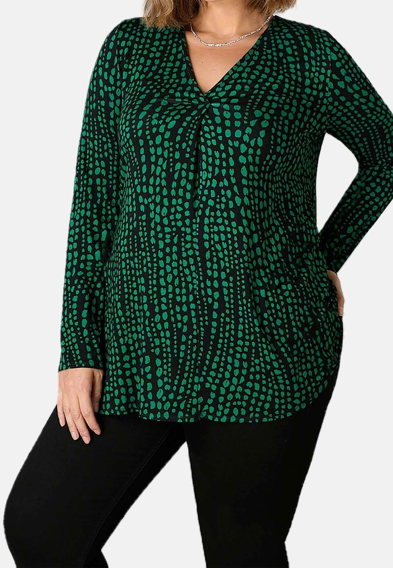 Frau trägt schwarzen Langarmpullover mit grünem unregelmäßigem Punktemuster, schwarze Hose und silberne Halskette, steht mit einer Hand in der Hüfte.