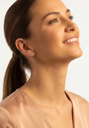 Giovane donna con capelli castani raccolti in una coda sorride, indossa una maglia marrone chiaro e piccoli orecchini a forma di stella, guarda leggermente verso l'alto.