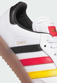Hvid Adidas Samba sneaker med sorte, røde og gule striber, gyldent "SAMBA" tekst og gennemsigtig gummisål, vist i close-up detaljer.