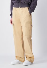 Beige cargobroek van katoen, met twee zijzakken en twee grote opgestikte zakken, gecombineerd met witte sneakers en een grijze sweatshirt.