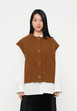 Bruine wollen vest met een ronde hals, grote armsgaten, knopen aan de voorkant en zijzakken, gelaagd over een wit shirt. Smooth textuur.