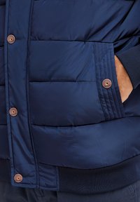 Marineblå dunjakke med kviltet mønster, der har metalknapdetaljer og ribbede manchetter. Glat tekstur, hofte-længde design.