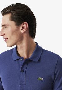 Polo de color azul marino con tejido texturizado, con tapeta de dos botones y logo de cocodrilo verde en el lado izquierdo del pecho. Mangas cortas y cuello clásico.