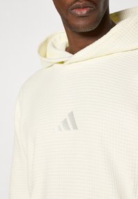 Sweatshirt com capuz amarelo claro, apresentando um padrão texturizado de waffle e um pequeno logótipo cinza da Adidas no peito. Capuz ajustado com cordões.
