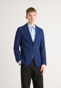 Blazer v modrem karirastem vzorcu z notnimi lapelami, zapiranjem na en gumb, stranskimi žepi, v kombinaciji s svetlobludem srajco s ovratnikom in črnimi hlačami.