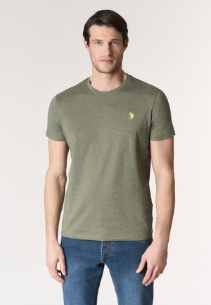 Uomo con t-shirt a maniche corte verde oliva con piccolo logo giallo sul petto e jeans blu, in piedi contro uno sfondo chiaro uniforme.