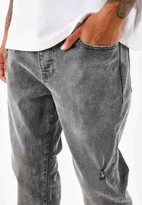 Jeans de mezclilla gris con un acabado ligeramente deslavado, que presentan un parche desgastado cerca del bolsillo y un diseño de pierna recta.