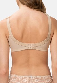 Beige rintaliivi, jossa on säädettävätolkaimet, läpinäkyvä takaosa ja monihakasuljenta. Herkkä tikkaus reunoilla lisää tekstuurillista yksityiskohtaa.