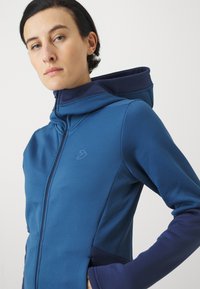 Giacca con cappuccio in blu profondo e navy, realizzata in tessuto elastico. Presenta una chiusura con zip, un logo discreto e un design aderente con maniche lunghe.