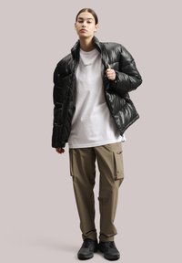 Melnas polsterētās jakas ar augstu apkakli, kas uzvilkta virs baltā T-kreklā un bēšajās cargo biksēs. Melnas sporta apavus papildina kopējo apģērbu.