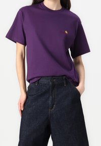 Persoon in een paarse korteknotted t-shirt met een klein logo op de borst en donkerblauwe hoge spijkerbroek, handen in de zakken.