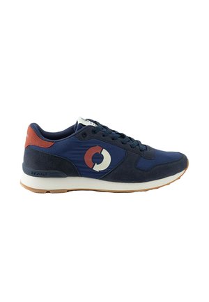 Zapatilla deportiva azul marino con logo circular blanco y marrón, acento marrón en el talón, borde de suela negro y entresuela blanca, vista lateral.