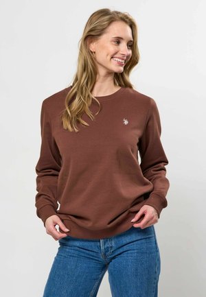 Jeune femme souriante, portant un sweat-shirt marron avec un petit logo blanc et un jean bleu, les mains ajustant l'ourlet du sweat-shirt.