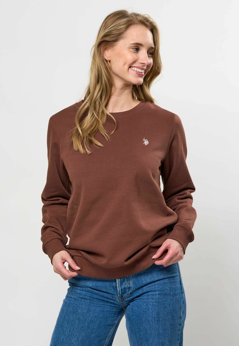 Jeune femme souriante, portant un sweat-shirt marron avec un petit logo blanc et un jean bleu, les mains ajustant l'ourlet du sweat-shirt.
