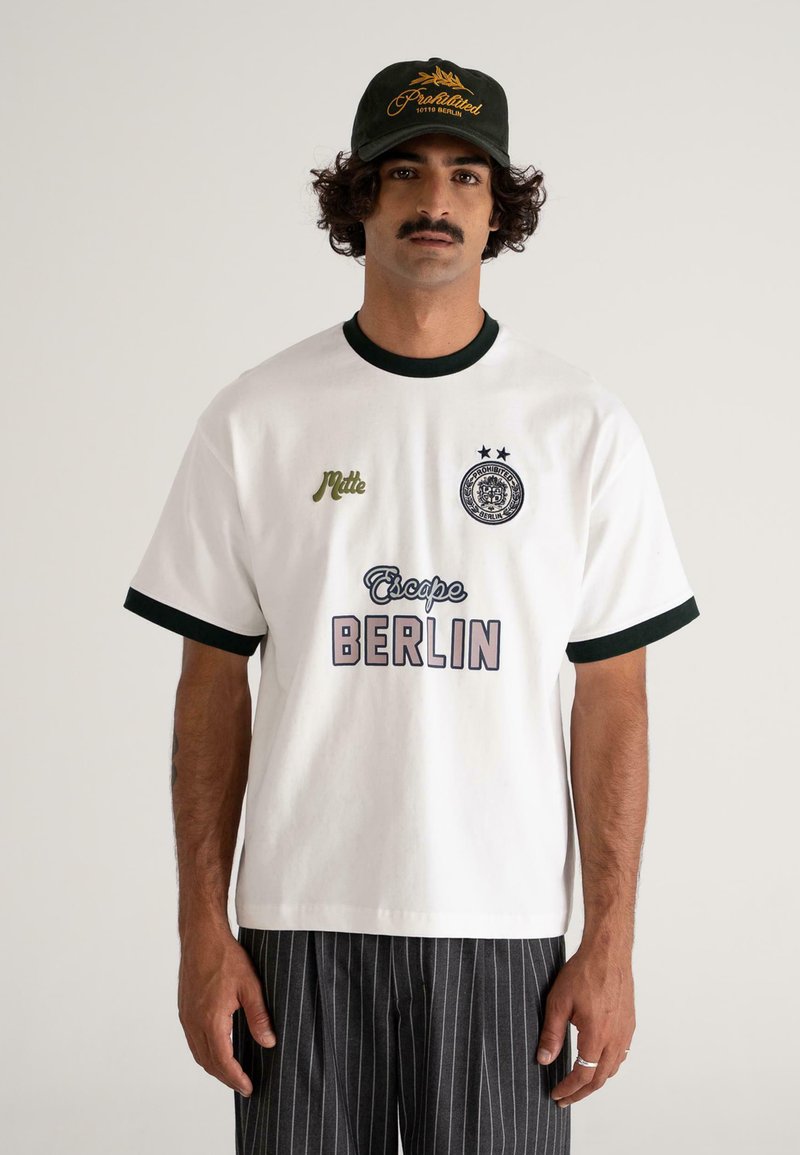 Bărbat cu mustață, purtând o șapcă neagră și un tricou alb cu margini întunecate, având inscripția "Escape BERLIN" și pantaloni dungati negri, stând în fața unui fundal simplu.
