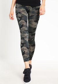 Persona che cammina indossando leggings con motivo mimetico, top nero e sneakers nere con suola bianca su sfondo bianco.