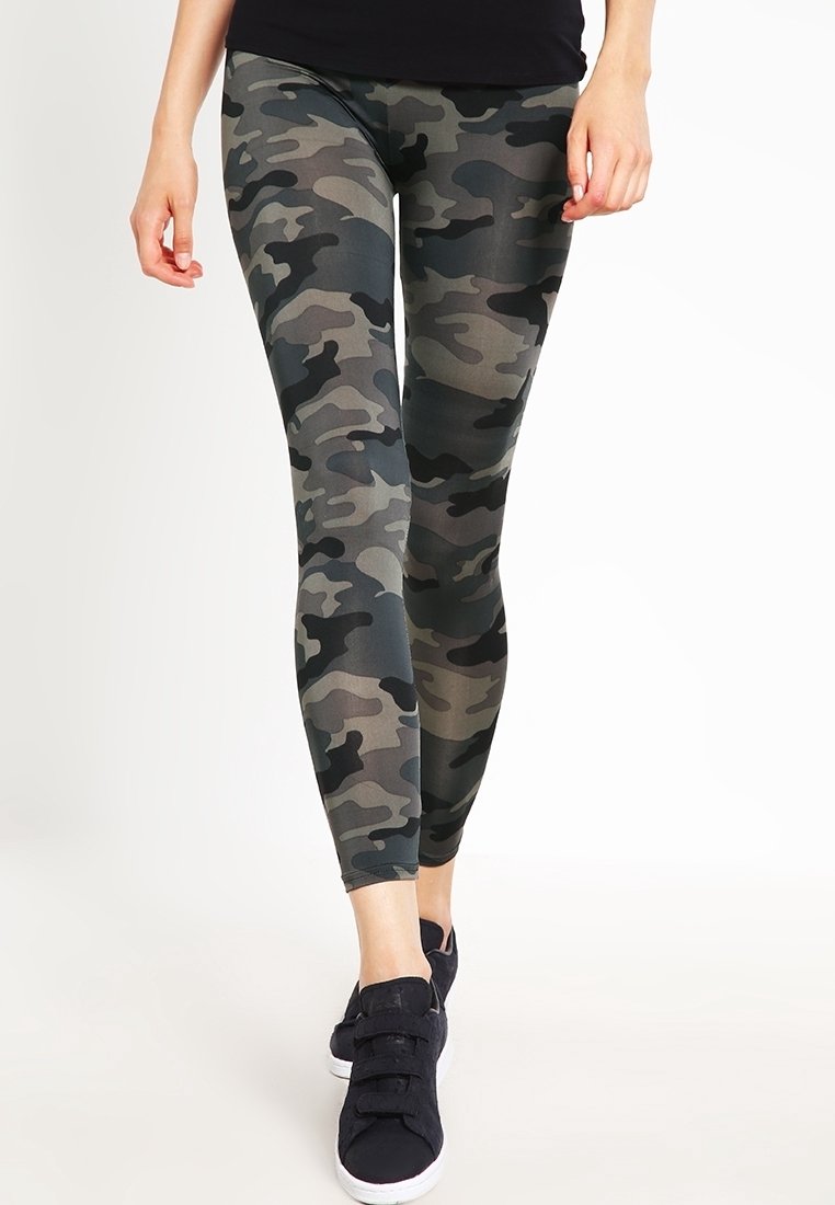 Persona che cammina indossando leggings con motivo mimetico, top nero e sneakers nere con suola bianca su sfondo bianco.