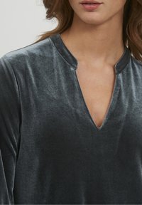 Mörkgrå sammetstopp med V-ringning och lång ärm. Tyget har en slät yta och en subtil lyster som reflekterar ljus.