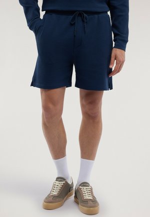 Hombre vistiendo pantalones cortos texturizados color azul marino con cordón, calcetines blancos hasta el tobillo y zapatillas casuales marrones, de pie contra un fondo liso.