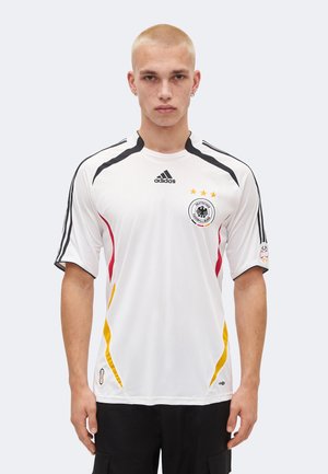 Jeune homme aux cheveux blonds courts portant un maillot de football blanc de l'Allemagne avec des accents noirs, rouges et jaunes, et le logo Adidas.