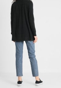 Svart långärmad kofta med en avslappnad passform, kombinerad med ljusblå korta jeans och svarta sneakers, mot en ljus bakgrund.