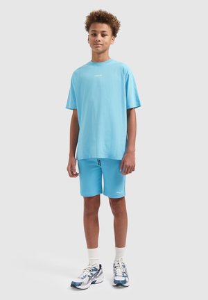 Tienerjongen staat en draagt een lichtblauw oversized t-shirt, bijpassende shorts, witte sokken en witte en blauwe hardloopschoenen.