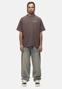 Brown oversized t-shirt met een ronde hals, vergezeld van lichtblauwe jeans en zwarte schoenen met witte accenten. Eenvoudig logo op het shirt.