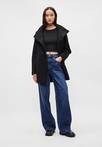 Čierny dvojradový kabát s veľkým golierom, nosený cez skrátený čierny top. Skombinovaný s vysoko pásovými širokými modrými džínsami a čiernymi čižmami.