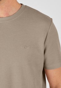 Camiseta de rayas beige con mangas cortas, que presenta un escote redondo y un pequeño logo en relieve en el pecho. Textura de tela de algodón visible.