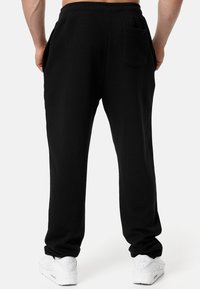 Black sweatpants made of soft fabric, featuring a back pocket and tapered legs, paired with white athletic sneakers on a neutral background.