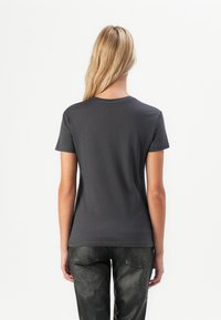 Blonde woman wearing a plain dark gray short-sleeve T-shirt and black textured pants, standing facing away against a white background.