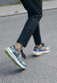 Scarpe sportive con un motivo floreale colorato in blu, rosa e verde; presentano una suola spessa e lacci neri, indossate con pantaloni neri.