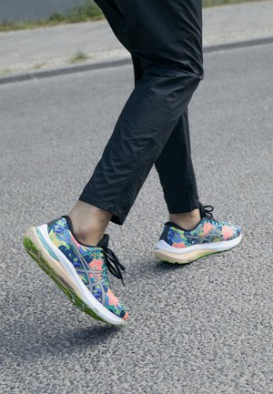Scarpe sportive con un motivo floreale colorato in blu, rosa e verde; presentano una suola spessa e lacci neri, indossate con pantaloni neri.