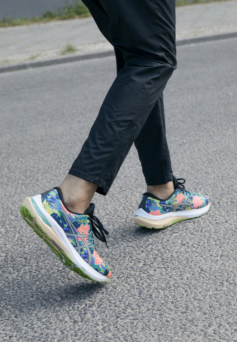 Scarpe sportive con un motivo floreale colorato in blu, rosa e verde; presentano una suola spessa e lacci neri, indossate con pantaloni neri.