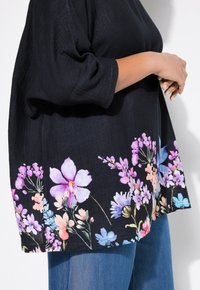 Schwarze oversized Bluse mit lebhaften floralen Mustern in Pink, Lila und Blau am Saum, aus weichem, strukturiertem Stoff und mit kurzen Ärmeln.