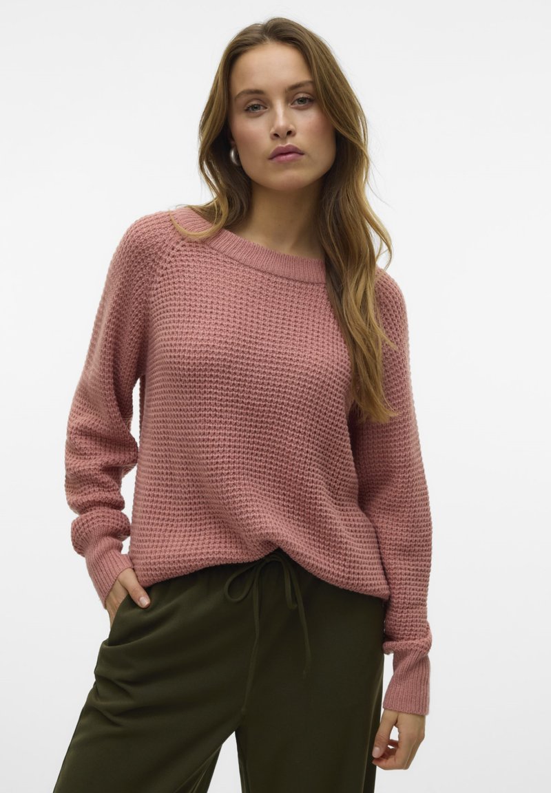 Pull en laine rose avec un motif texturé, col large et manches fuselées, associé à un pantalon à cordon de serrage vert foncé.
