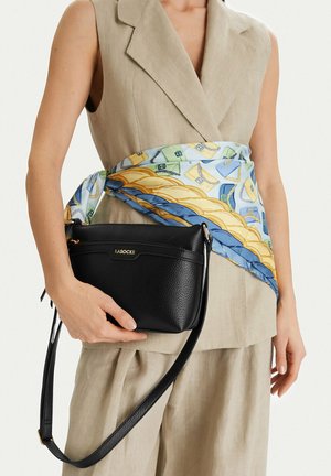 Mujer con traje sin mangas beige sosteniendo un bolso cruzado negro de Lasocki con una bufanda de colores y estampado atada alrededor de su cintura.