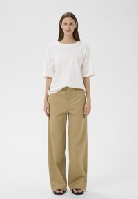 Jeune femme debout, vue de face, portant une chemise blanche à manches courtes oversize, un pantalon beige à jambes larges et des sandales noires.