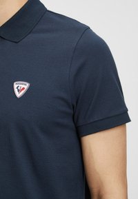 Námořnická modrá polo košile z texturované látky s krátkým rukávem, která má trojúhelníkovou nášivku loga Rossignol na levé části hrudníku.