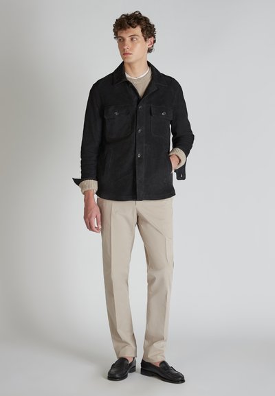 Giacca nera in suede con due tasche sul petto, chiusura a bottoni e un ampio colletto, abbinata a pantaloni beige e mocassini neri.