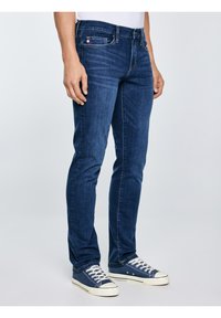 Személy sötétkék, testhezálló farmernadrágban és fehér fűzős tengerészkék vászon tornacipőben, sima világos háttér előtt állva.