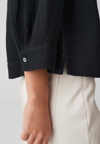 Zwarte, gestructureerde shirt met een geblokt patroon; met een manchet met een knoopdetail, gedragen over lichte broek.