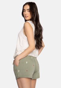 Olijfgroene shorts met cactus- en hartpatronen, zijzakken en een relaxed fit, gecombineerd met een witte mouwloze top.